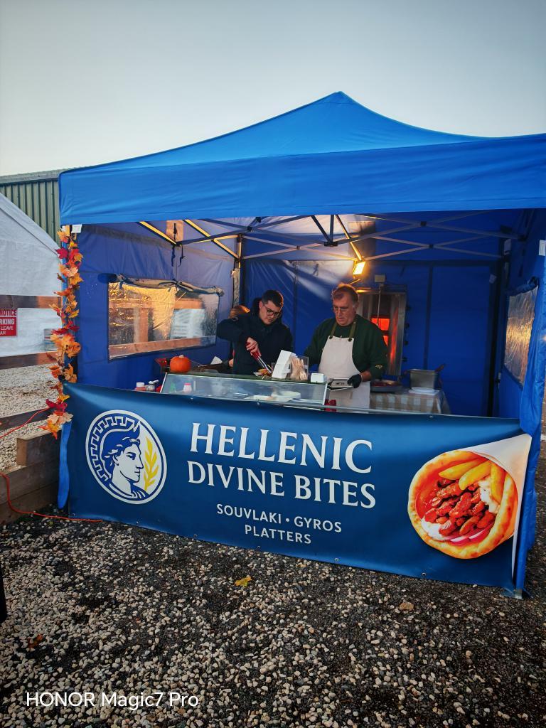 Two people work at a food stall under a blue tent labeled "Hellenic Divine Bites," serving souvlaki, gyros, and platters. Various kitchen items are on the counter.