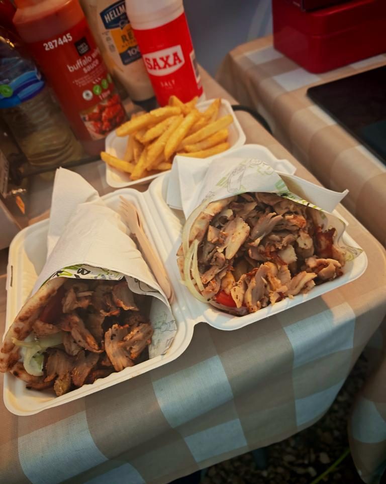 Two open takeaway boxes with chicken gyros wrapped in pita bread and a portion of French fries on a table, surrounded by various condiments.